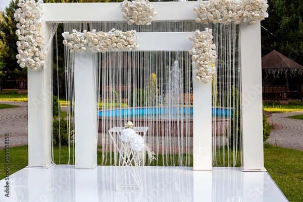 Obraz Outdoor wedding setup featuring a modern white arch adorned with cascading white roses and ribbons, positioned in front of a fountain in a scenic garden setting