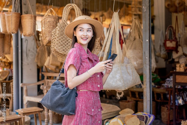 Fototapeta Asian woman tourist using phone at traditional handicraft store in Chiang Mai. Gen Z female exploring local market and sharing moments on social media.