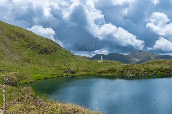 Fototapeta Capra Lake in the Fagaras Mountains, Romania. Cloudy sky over a calm alpine lake and green mountain slopes in summer.
