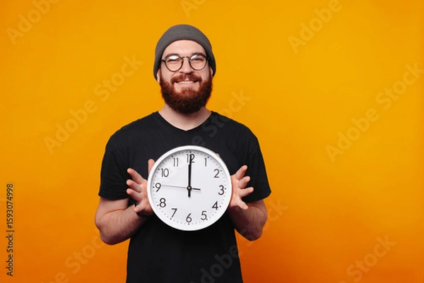 Fototapeta A joyful and cheerful man is happily holding a clock in front of a bright and vibrant orange background