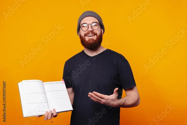 Obraz A smiling man holding a notebook, ready to share ideas or lessons against a vibrant yellow backdrop.