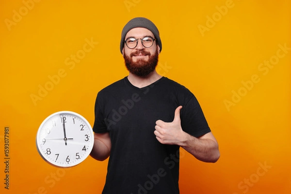 Obraz A cheerful young man gives a thumbs up while holding a clock against a bright yellow background, exuding joy
