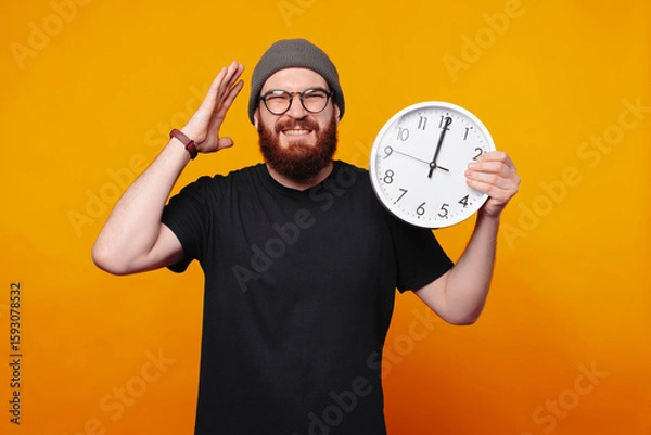Obraz A cheerful, smiling man holding a clock, showing urgency or surprise in a bright and lively setting