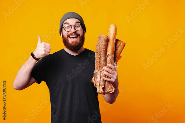 Obraz A joyful man expresses his enthusiasm as he proudly holds up a loaf of fresh bread while giving a thumbsup gesture