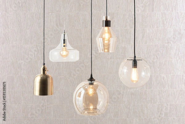 Fototapeta Assorted hanging pendant lights in sculptural clear glass and brushed metal finishes with exposed filament bulbs, set against a textured beige wall, evoking a modern eclectic style with warm ambiance