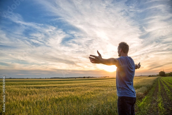 Fototapeta Man standing in an open field at sunset with open arms - embracing nature
