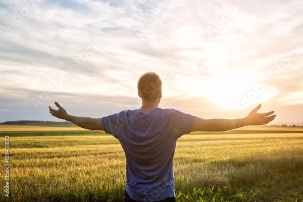 Fototapeta Man standing in an open field at sunset with open arms - embracing nature