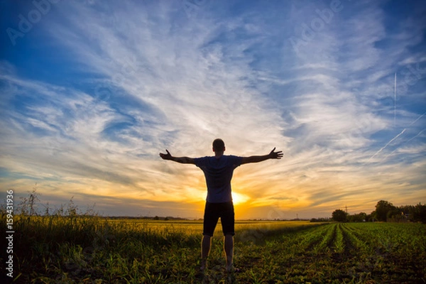 Fototapeta Man standing in an open field at sunset with open arms - embracing nature