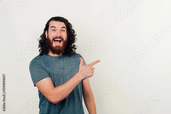 Obraz A cheerful and happy man with curly hair is energetically pointing at an empty space, radiating positivity