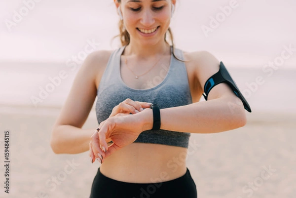 Obraz A smiling woman checks her fitness tracker while exercising on the beach, promoting a healthy lifestyle.