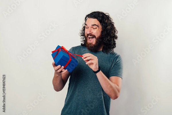 Obraz A joyful man with curly hair and a beard unwraps a blue present, showcasing surprise and excitement.
