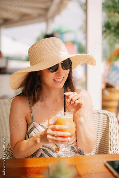 Obraz A stylish woman in a widebrimmed hat relaxes, sipping a colorful cocktail at a beachside caf