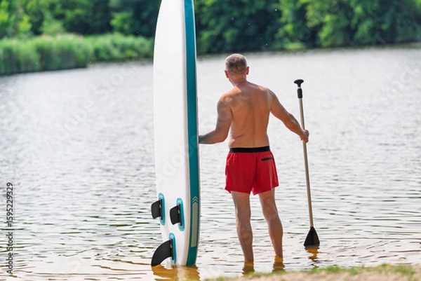 Fototapeta A man stands by the river with a paddle and a SUP board.