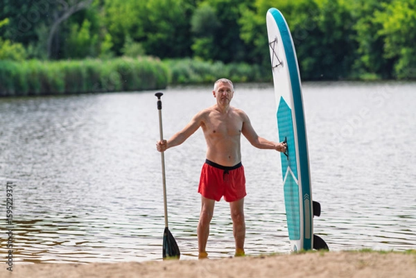 Fototapeta Middle aged man by the river with a SUP board