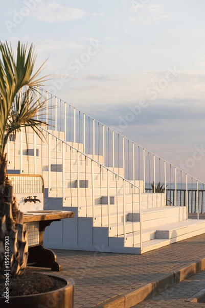 Fototapeta Staircase leading to a seaside restaurant at sunset, with a view of the calm ocean, a sailboat in the distance, and tropical palm leaves in the foreground.