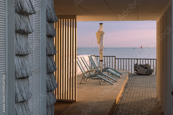 Fototapeta Seaside terrace with empty lounge chairs and a closed umbrella at sunset, overlooking calm ocean water and distant sailboats.