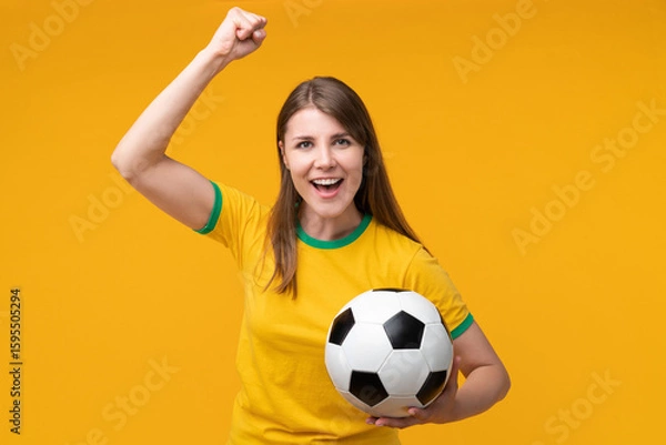 Obraz Happy young woman in yellow shirt cheering with raised fist and holding soccer ball