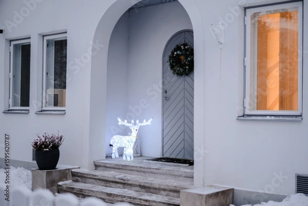Fototapeta A charming house entrance adorned with festive winter decorations, featuring an illuminated glowing reindeer standing on the snow-covered steps