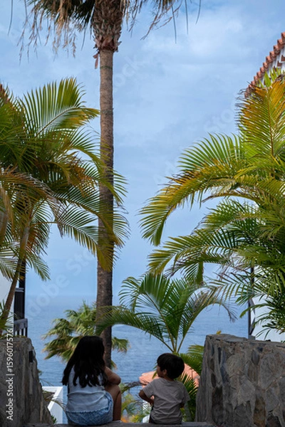 Fototapeta Two children are sitting on a stone staircase overlooking the ocean. The scene is peaceful and serene, with palm trees in the background and the sound of the waves in the distance