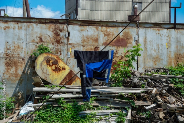 Obraz Clothes are suspended on a line in an overgrown area with rust-stained metal surfaces and piles of discarded materials