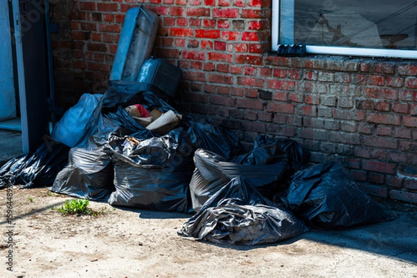 Obraz Several black trash bags stuffed with waste are stacked against a red brick wall in a commercial setting, highlighting urban litter issues