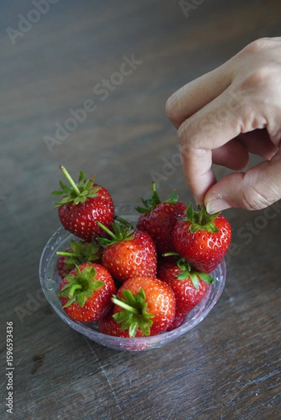 Fototapeta closeup hands are picking strawberries. Many fresh strawberries in a clear plastic glass lid on a wooden table background, fruit, food, decor, object, copy space