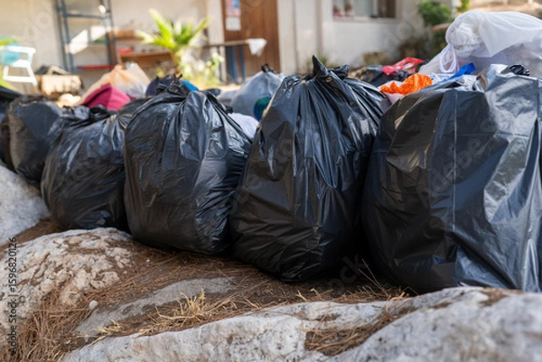 Fototapeta Black trash bags are stacked along a rocky area, ready for collection. Various discarded items can be seen, reflecting efforts to manage waste responsibly.