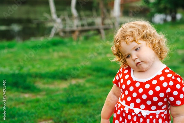 Fototapeta A young child wearing a bright red dress with white polka dots stands with a thoughtful expression in a grassy area The sun casts a warm light as she gazes down