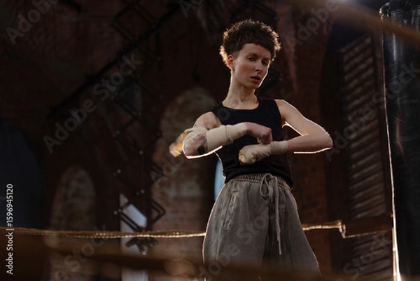 Fototapeta A woman in a boxing ring, wrapping an elastic bandage around her arms, preparing for boxing . The photo is in a cinematic style, the light gives the mood to a sports motivational story about self