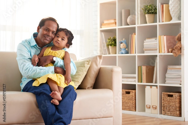Fototapeta Portrait of loving Indian father hugging his pretty little girl and posing for photography  in cozy living room