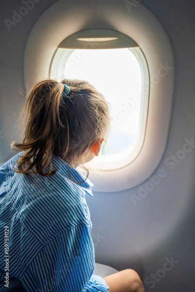 Fototapeta Little girl looking out the airplane window during a flight. Wonder, travel experience, and childhood curiosity in the air.