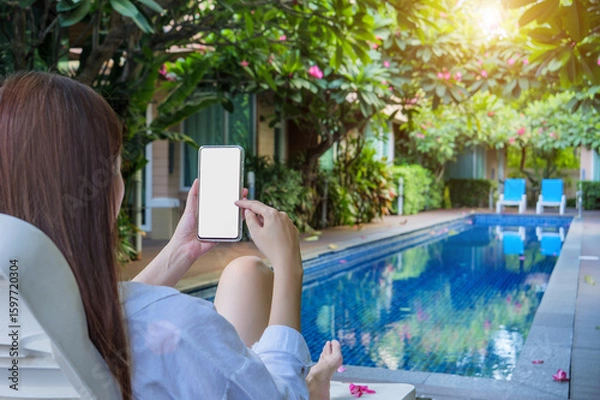 Fototapeta a woman using finger for touch on phone mockup with blank white screen with copy space, relaxing by the poolside on vacation day at hotel resort.