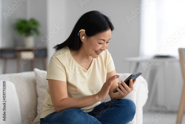 Obraz A forty-year-old deaf asian  woman with a hearing aid, with short black hair, stubble, wearing a light yellow t-shirt and jeans, sits on a white fabric sofa holding a smartphone. The living room