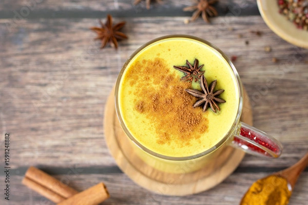 Fototapeta Golden milk (turmeric latte drink) with cinnamon and anise star on wooden table. Top view.