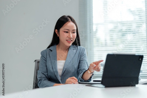 Fototapeta Young Asian businesswoman in suit having video call on tablet, pointing at screen and talking to client or coworker, working remotely in modern office