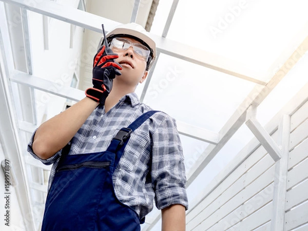 Fototapeta Confident Asian man civil engineer looking forward and talking on walkie talkie. Smart male foreman using radio communication equipment at construction site for cooperation with the worker team