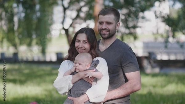Fototapeta A joyful family stands together in a park, with the mother holding their baby and the father smiling beside them, all basking in the warm sunshine.