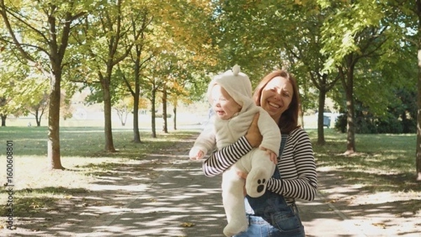 Fototapeta Smiling mother strolls through a park, carrying a baby in a cozy outfit while enjoying the warm sunlight filtering through vibrant autumn leaves.