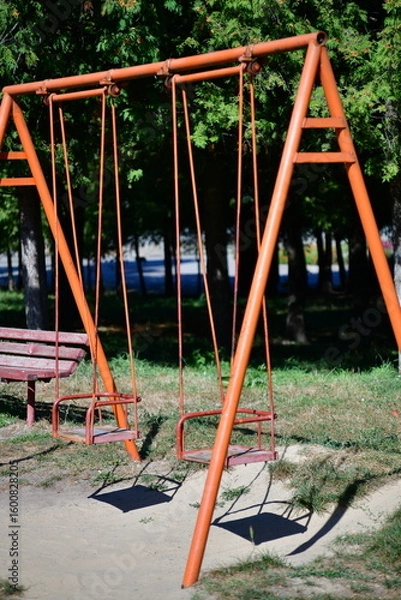 Fototapeta Swings in a quiet park surrounded by trees on a sunny day inviting children to play and enjoy outdoor activities