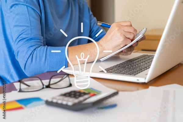 Fototapeta Businesswoman having new ideas and taking notes while working at her desk with a laptop and calculator, a light bulb graphic representing the concept of innovation and business intelligence