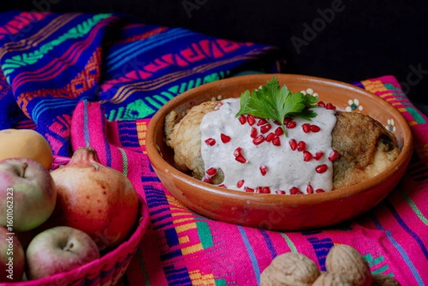 Fototapeta Classic Mexican chile en nogada served in a clay dish with fresh ingredients: apple, pear, walnut, and pomegranate over a vibrant textile background.
