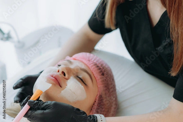 Fototapeta professional cosmetologist applies a creamy face mask on yiung woman face using a brush
