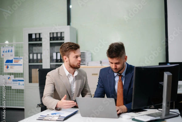 Fototapeta Professional businessmen collaborating in modern office, using laptop and tablet, discussing strategy, teamwork, and innovation.
