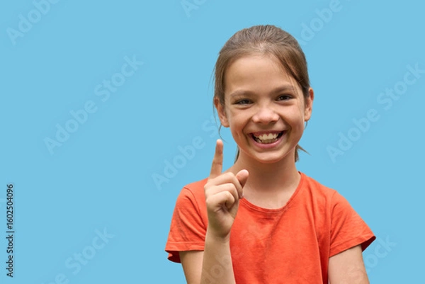 Fototapeta Cheerful White girl in her 10s smiling and pointing up with index finger on blue background. Concept of bright idea, childhood positivity, and educational communication