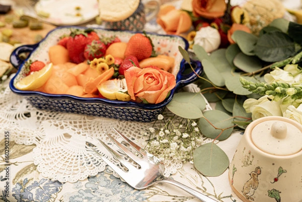 Fototapeta ceramic dish filled with strawberries, melon cubes, lemon slices, and decorated with orange roses and flowers on a lace tablecloth for an outdoor garden party