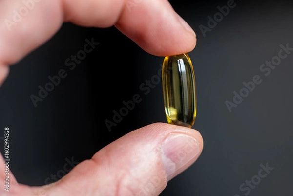 Fototapeta Yellow softgel capsule held between fingers on dark background. Close-up studio shot of transparent dietary supplement. Healthcare and pharmaceutical concept for design and banner