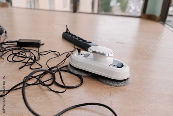 Fototapeta Window cleaning robot resting on floor, plugged into power strip, charging before window-washing operation at home, nobody. Modern window cleaning robot connected to chargers and power strip.