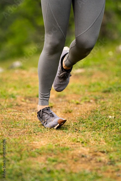 Fototapeta Woman in sportswear spraining ankle while running outdoors, vertical close-up of foot twisting on uneven grass surface in nature, risk of sports injury and leg trauma during outdoor activity