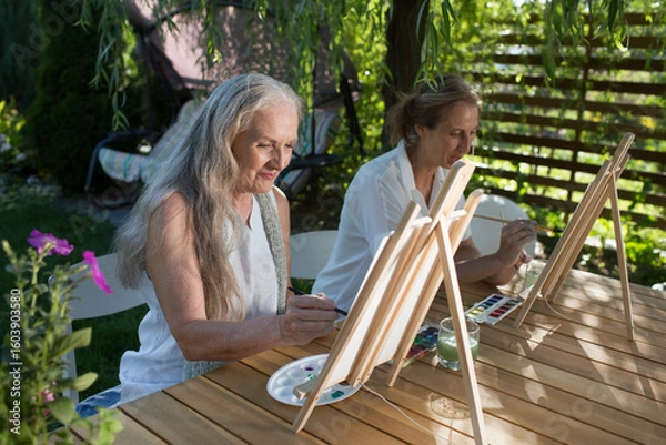 Fototapeta Two happy middle aged woman smiling while drawing watercolor as a recreational activity or therapy outdoors together with the group of retired women. KnowledgeForAll
