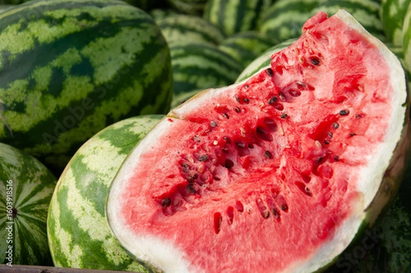 Fototapeta Close-up of a ripe watermelon cut in half, revealing juicy red flesh and black seeds, surrounded by whole green-striped watermelons. Fresh and vibrant image good for food advertising, summer concept